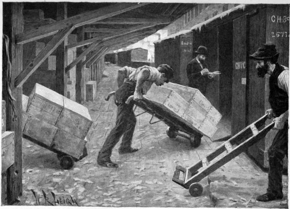 Three men are under cover on a loading dock.
There are box cars next to the loading dock.
One of the men is pushing a hand truck loaded with
crates into one of the boxcars.